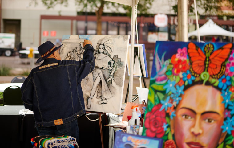 Photo of an artist painting on a canvas at an outdoor art event, with vibrant portrait artwork featuring flowers and butterflies displayed behind them.